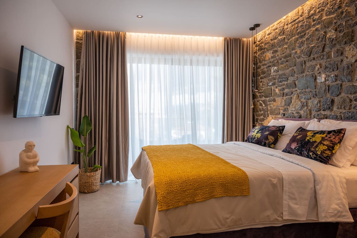 A modern bedroom with a stone wall, a large bed with white bedding and a yellow throw, a flat-screen TV on the wall, and a wooden desk with a plant.
