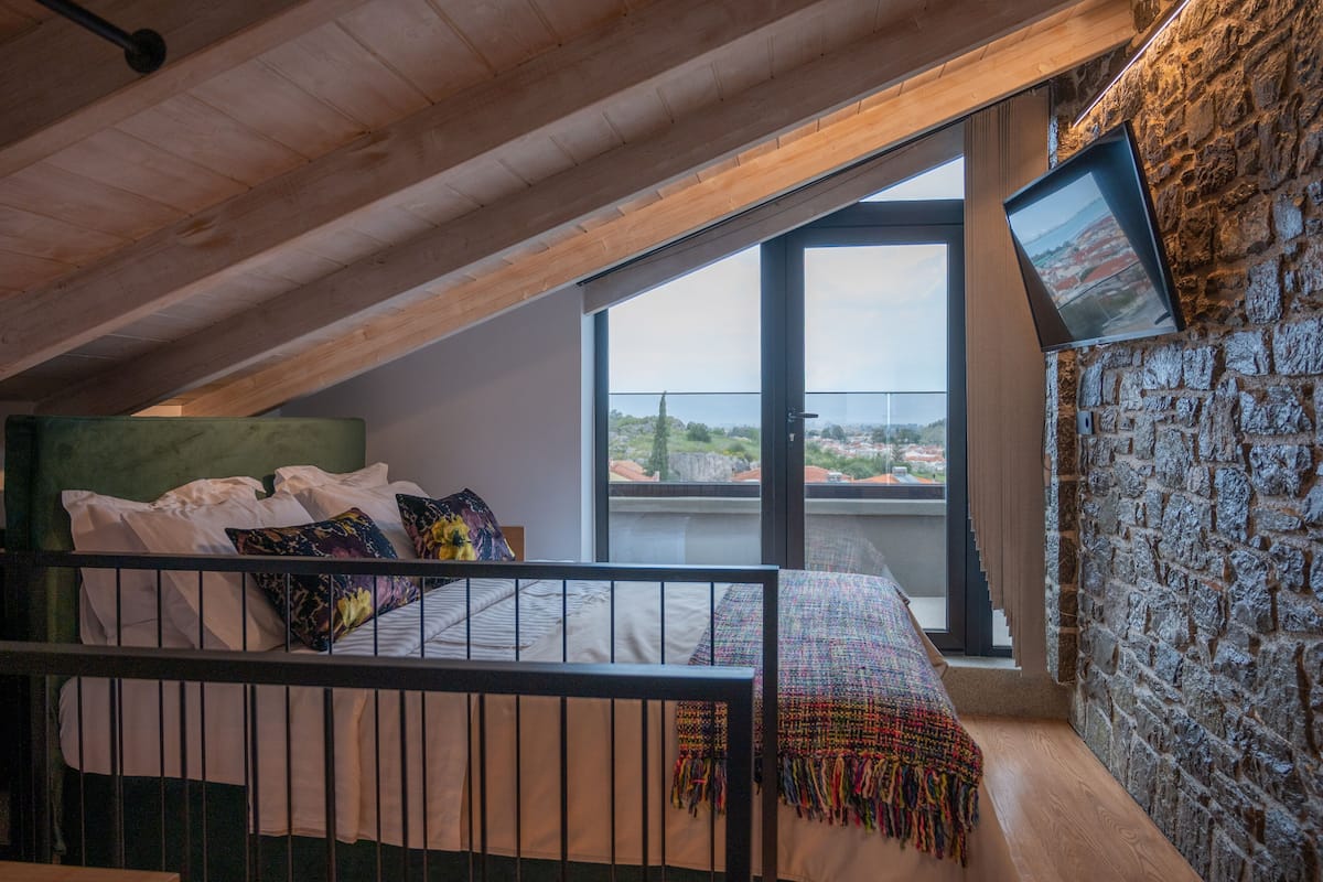A bedroom with a sloped ceiling, a large bed with pillows, a stone wall, and a TV mounted on the wall.