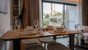 A dining table set with plates, bowls, glasses, and cutlery, with wicker chairs and a view of buildings through a window. - Opal Executive Suites (Nafplio)