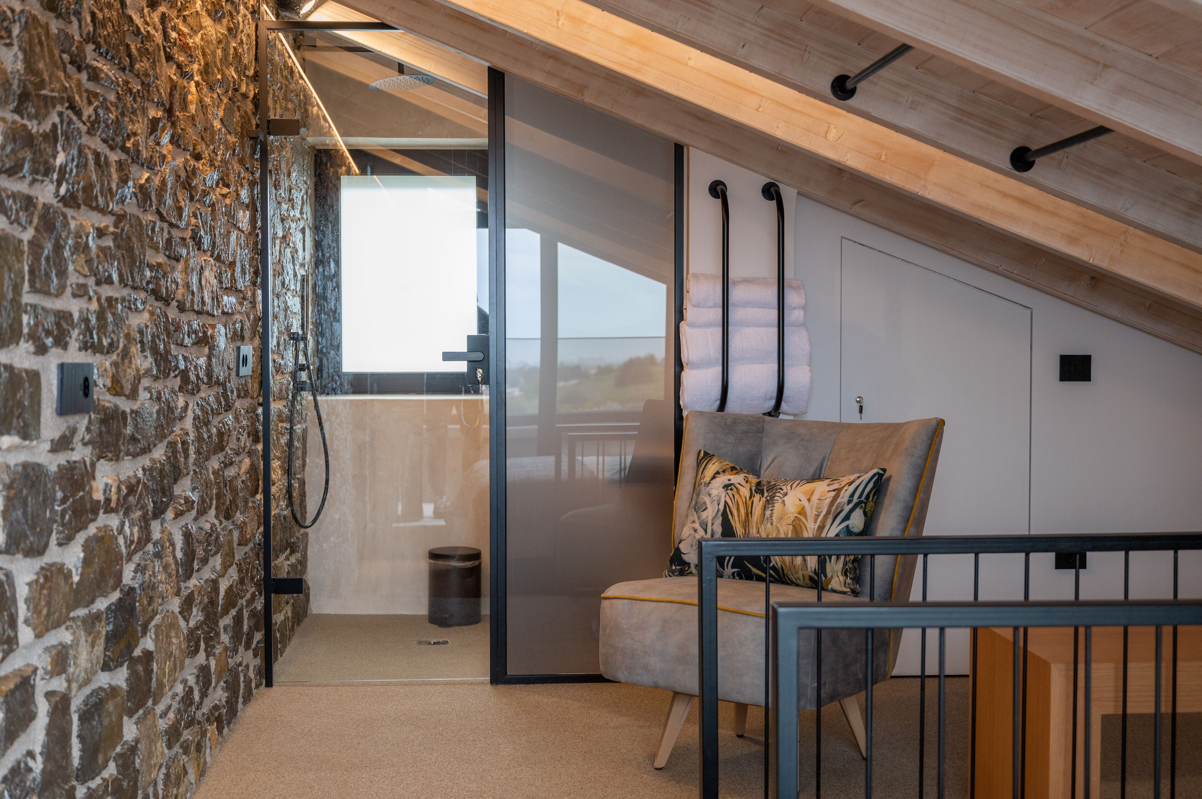 A cozy attic bedroom with a stone wall, a modern chair, and a view through a glass door.