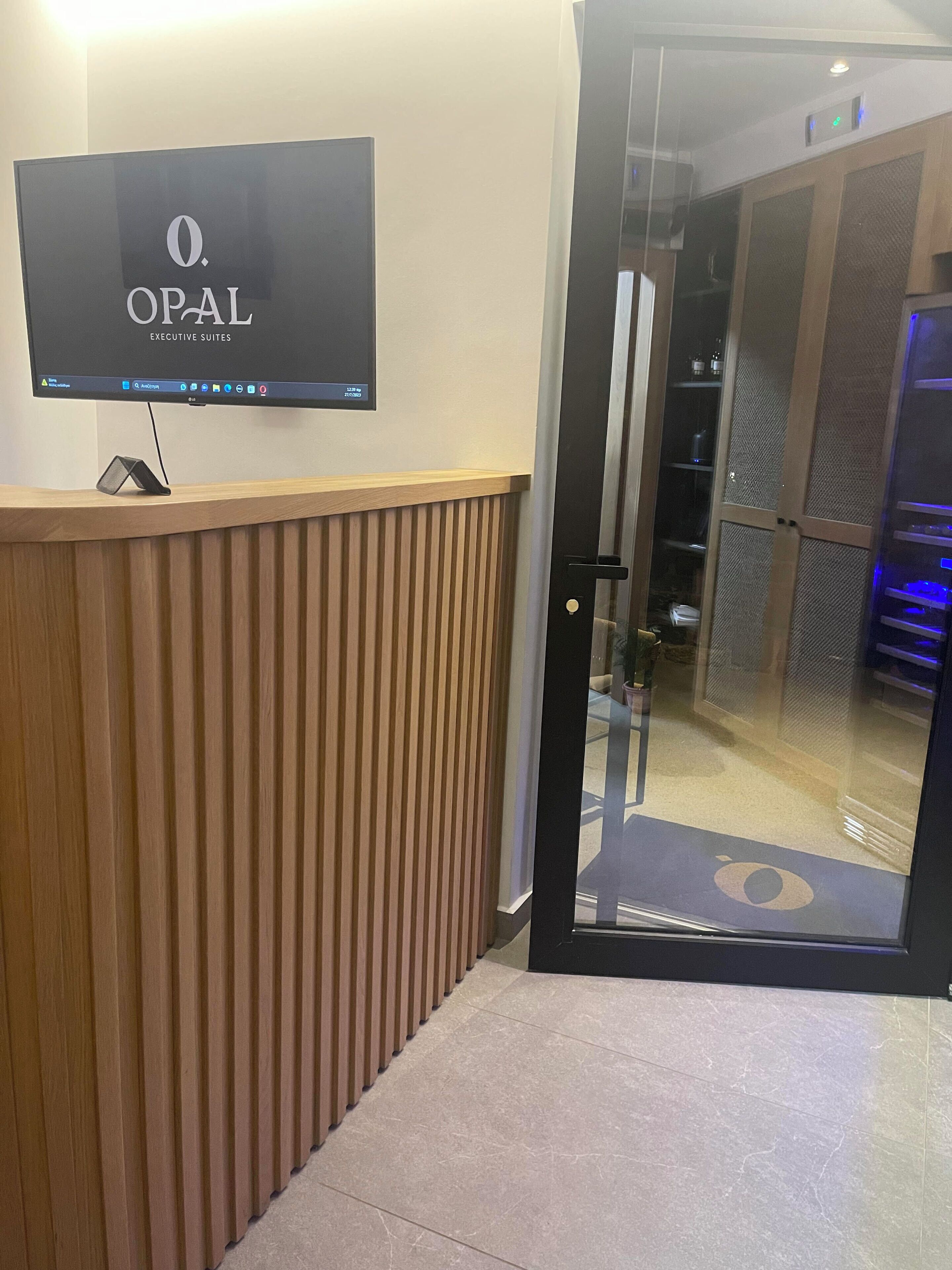 Una zona de recepción con un mostrador de madera, un televisor de pantalla plana que muestra 'OPAL EXECUTIVE SUITES' y una puerta de cristal que da a una sala con estanterías y una zona iluminada en azul.