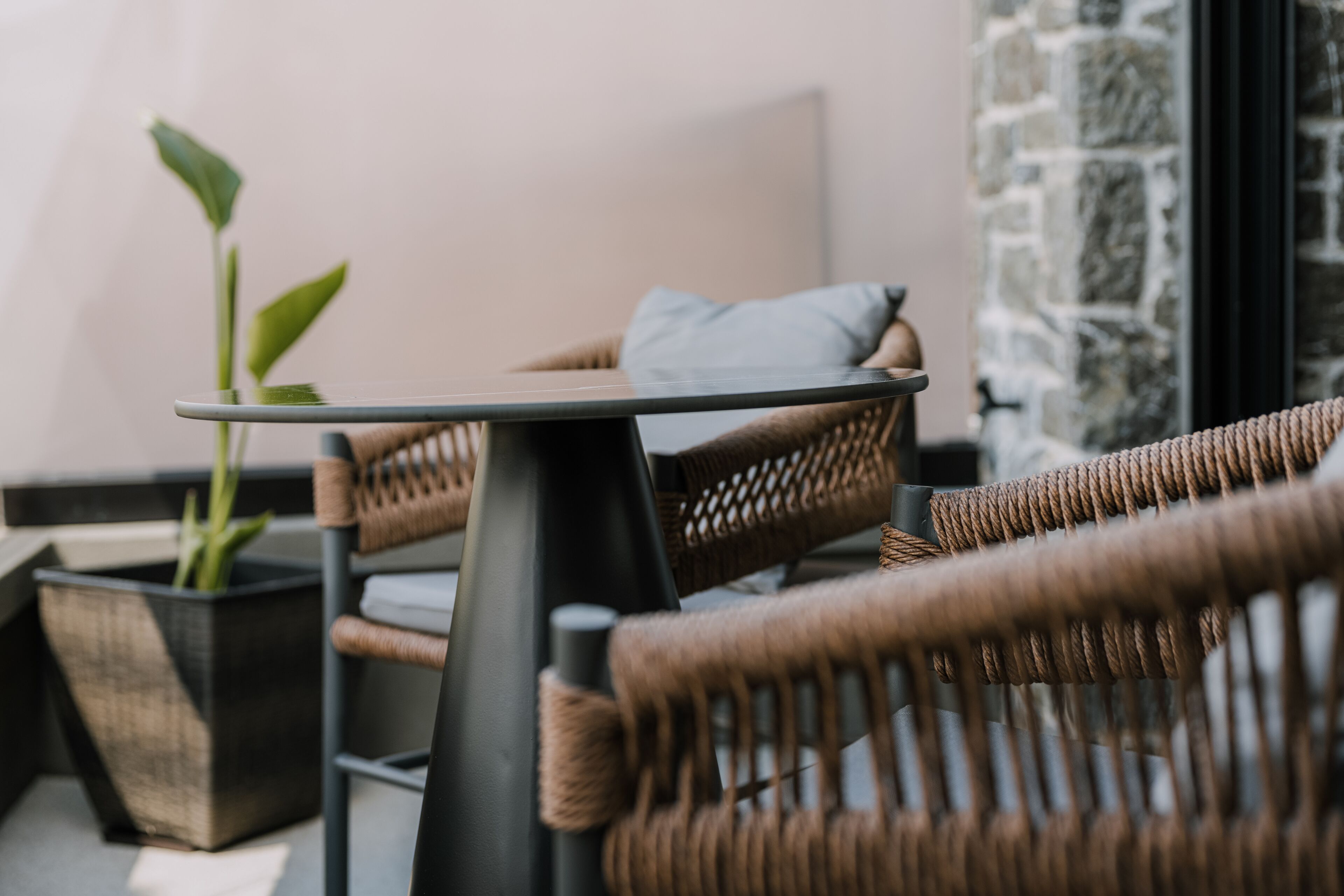 A wicker chair and table set with a potted plant, a stone wall, and a window in the background.