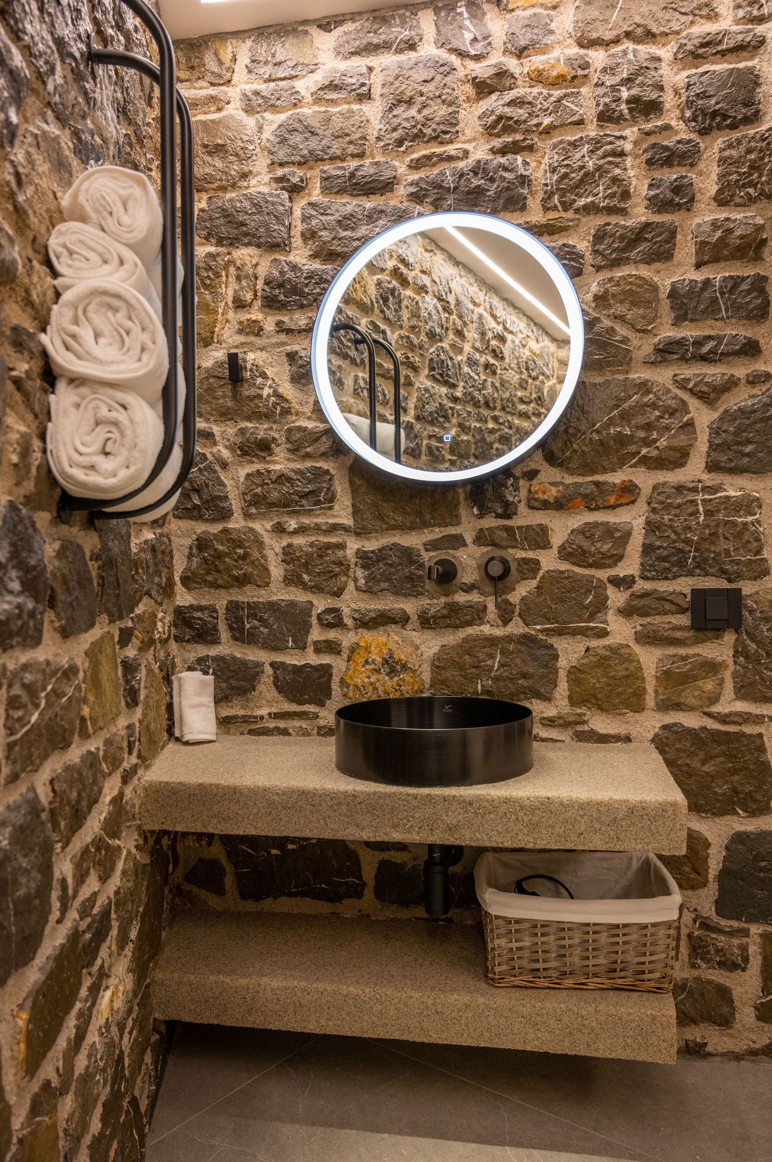 A stone-walled bathroom with a round mirror, a sink, and a towel rack with neatly rolled towels.