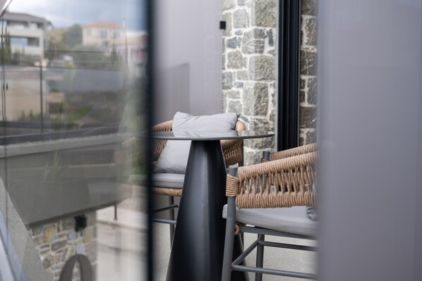 A balcony with wicker furniture and a glass table, overlooking a stone wall and buildings.