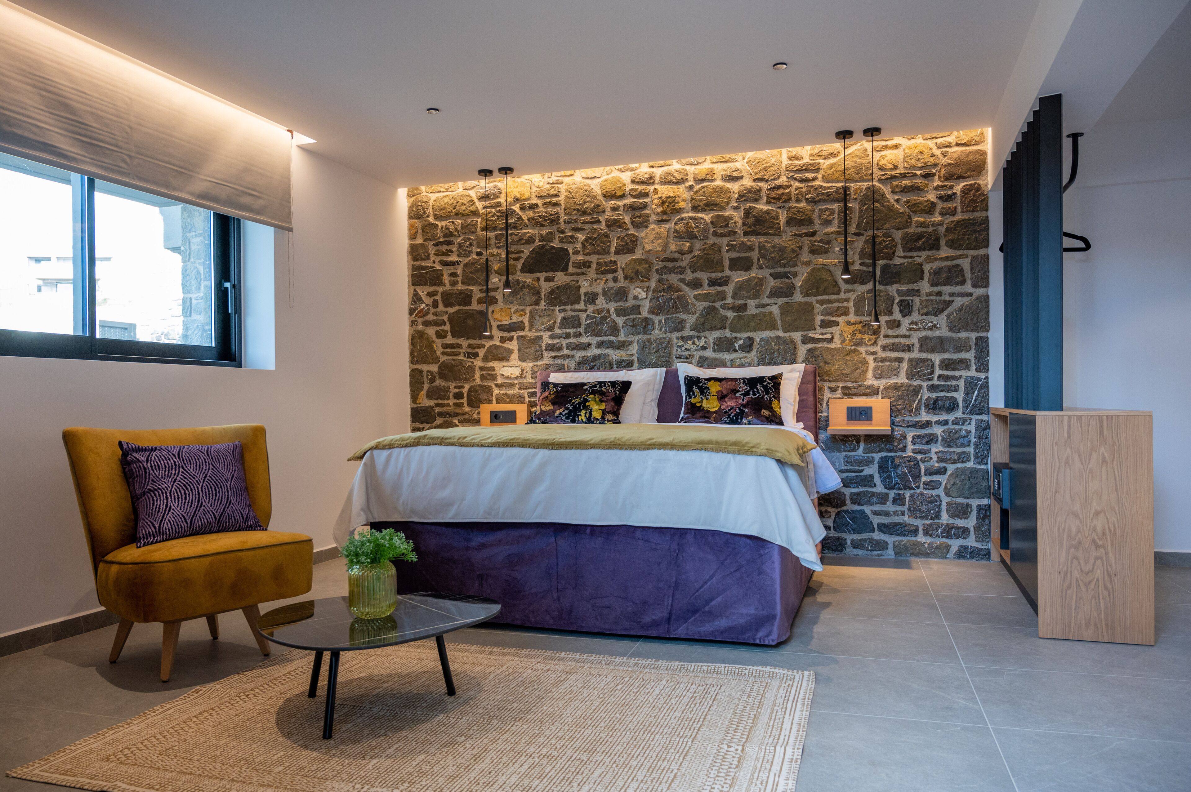 Ein modernes Schlafzimmer mit Steinwand, einem Bett mit weiß-lila Bettdecke, einem gelben Sessel und einem kleinen Tisch mit Pflanze.