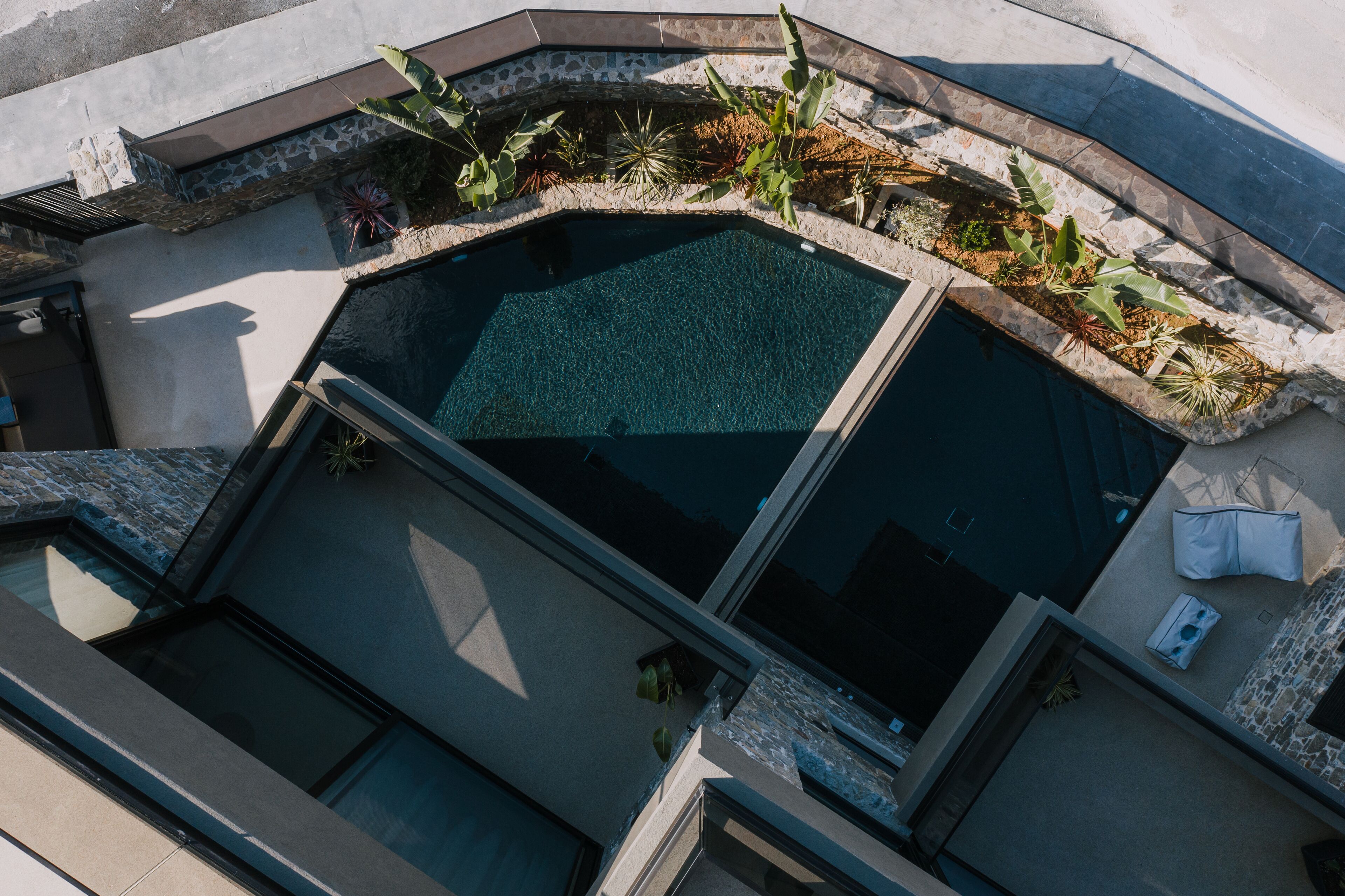 A rooftop pool with a stone border and surrounding plants, surrounded by a building with large windows and a stone facade.