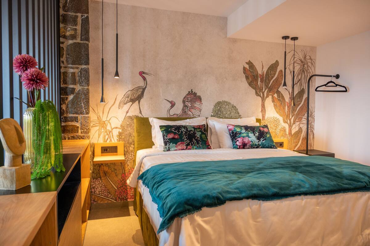 A modern bedroom with a stone wall, a bed with floral pillows, a teal throw blanket, and a hanging light fixture.