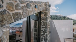 A stone wall with a glass door leading to a balcony with a view of a cityscape and a hill. - Opal Executive Suites (Nafplio)