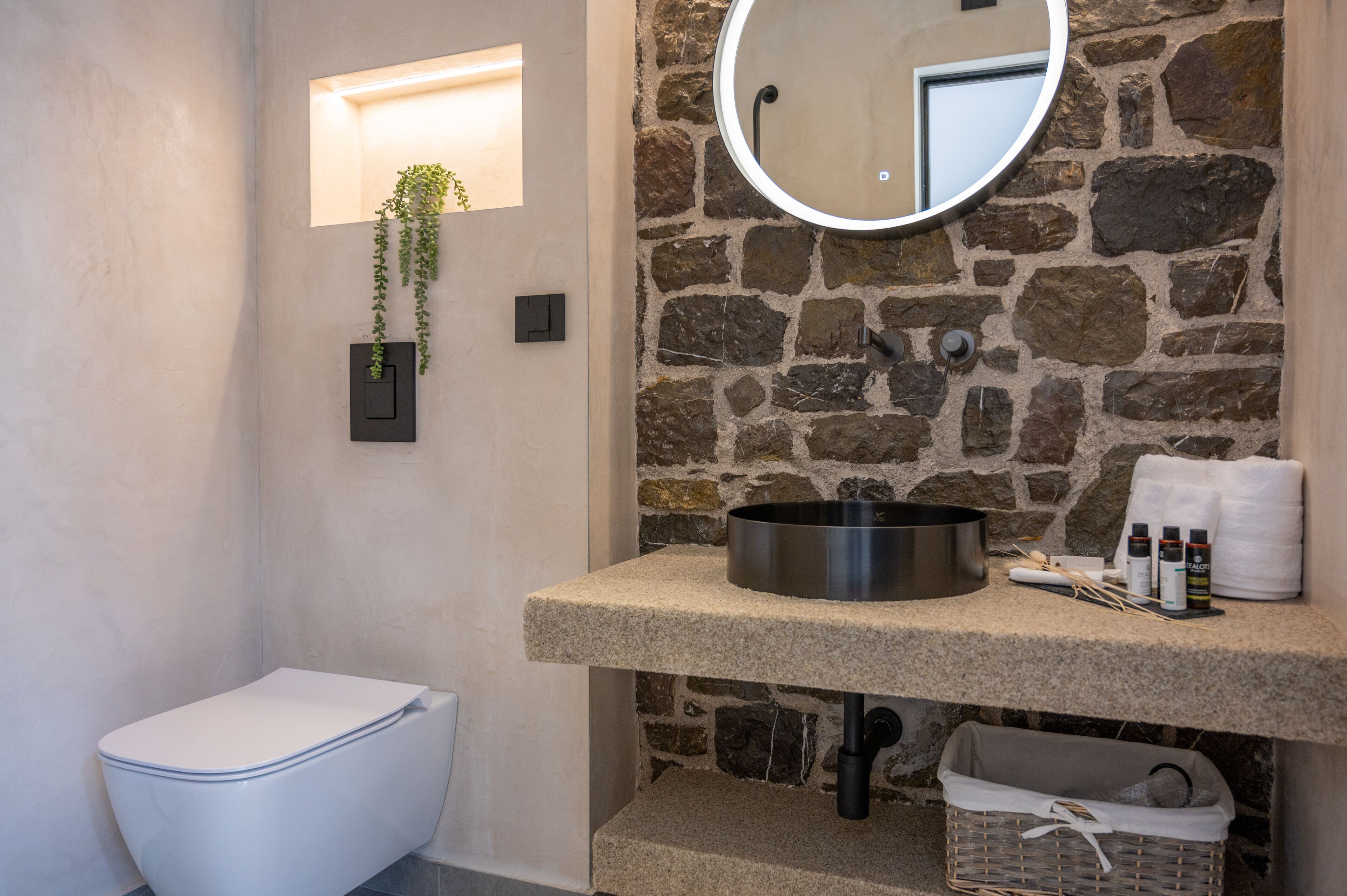 Un baño con pared de piedra, un espejo redondo, un lavabo con artículos de aseo y un inodoro.