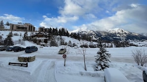 A snow-covered landscape with a mountain in the background, a road with parked cars, and buildings with a snow-covered roof.