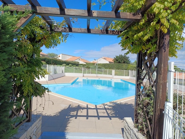A swimming pool with a wooden pergola overhead, surrounded by greenery and a white fence.