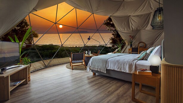 A tented accommodation with a bed, bedside table, lamp, and a view of the sunset over the sea. - Eco Dome Crete (Agios Nikolaos)