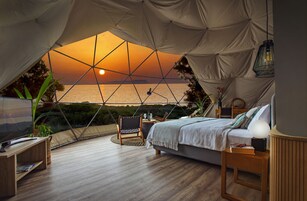 Alojamiento en carpa con cama, mesita de noche, lámpara y vistas al atardecer sobre el mar.