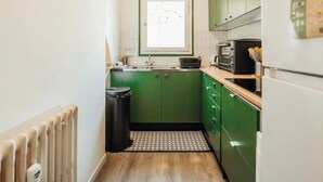 Eine kleine Küche mit grünen Schränken, einer schwarzen Mülltonne, einem Fenster und einem weißen Kühlschrank.