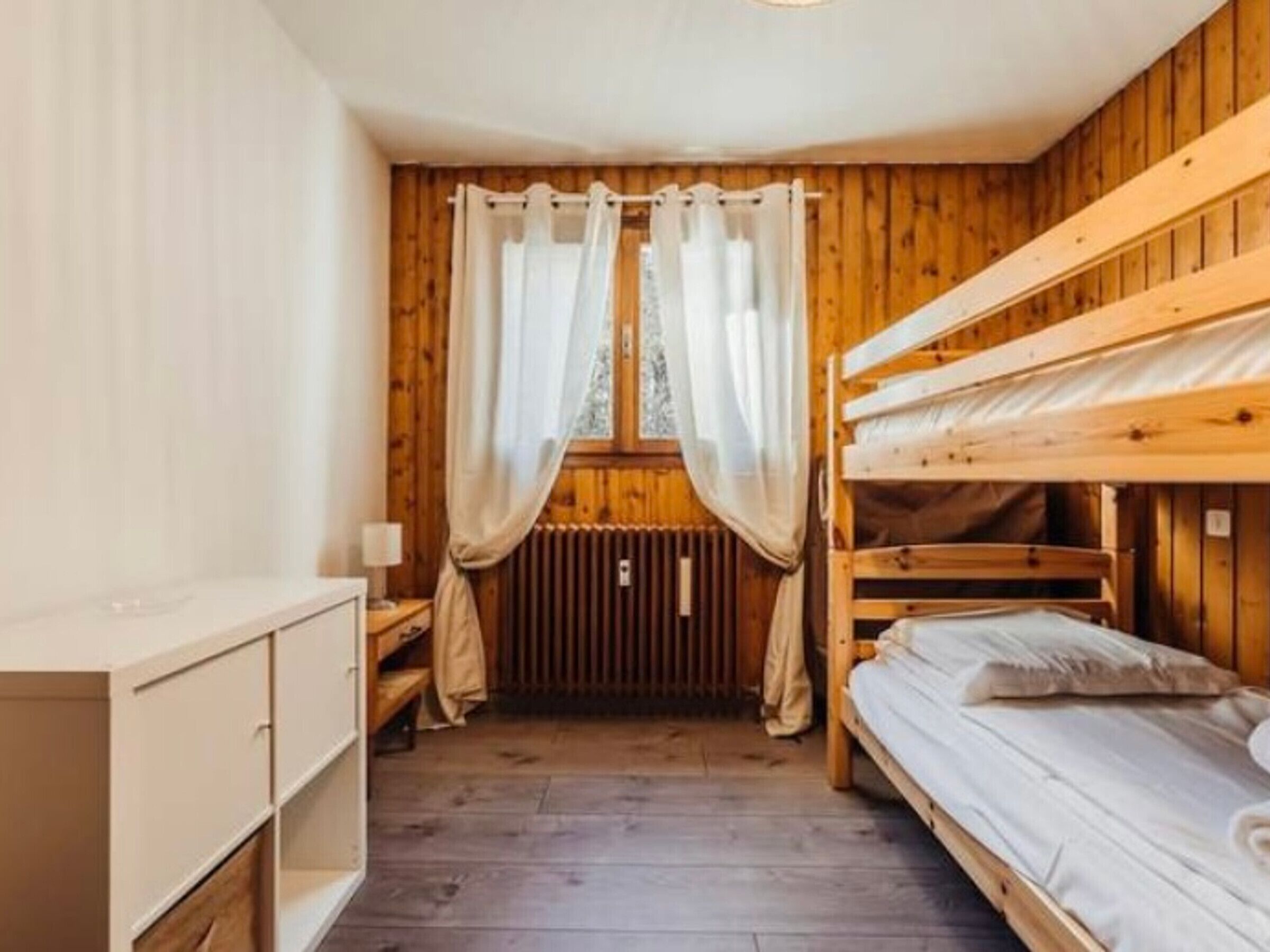 A cozy cabin room with a bunk bed, wooden walls, a window with curtains, and a radiator.