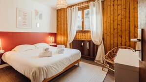 Ein Schlafzimmer mit Holzboden, einem Bett mit weißer Bettwäsche, einem Holz-Kopfteil, einer Pendelleuchte, einem Fenster mit Vorhängen und einer Heizung.