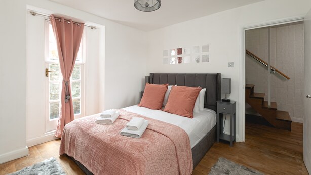 A bedroom with a bed, a nightstand, a lamp, a window with curtains, and a staircase.