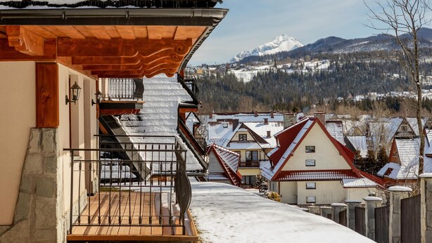 Ein mit Schnee bedeckter Balkon mit Holzgeländer, der auf ein Dorf mit traditionellen Häusern und einen Berg im Hintergrund blickt.