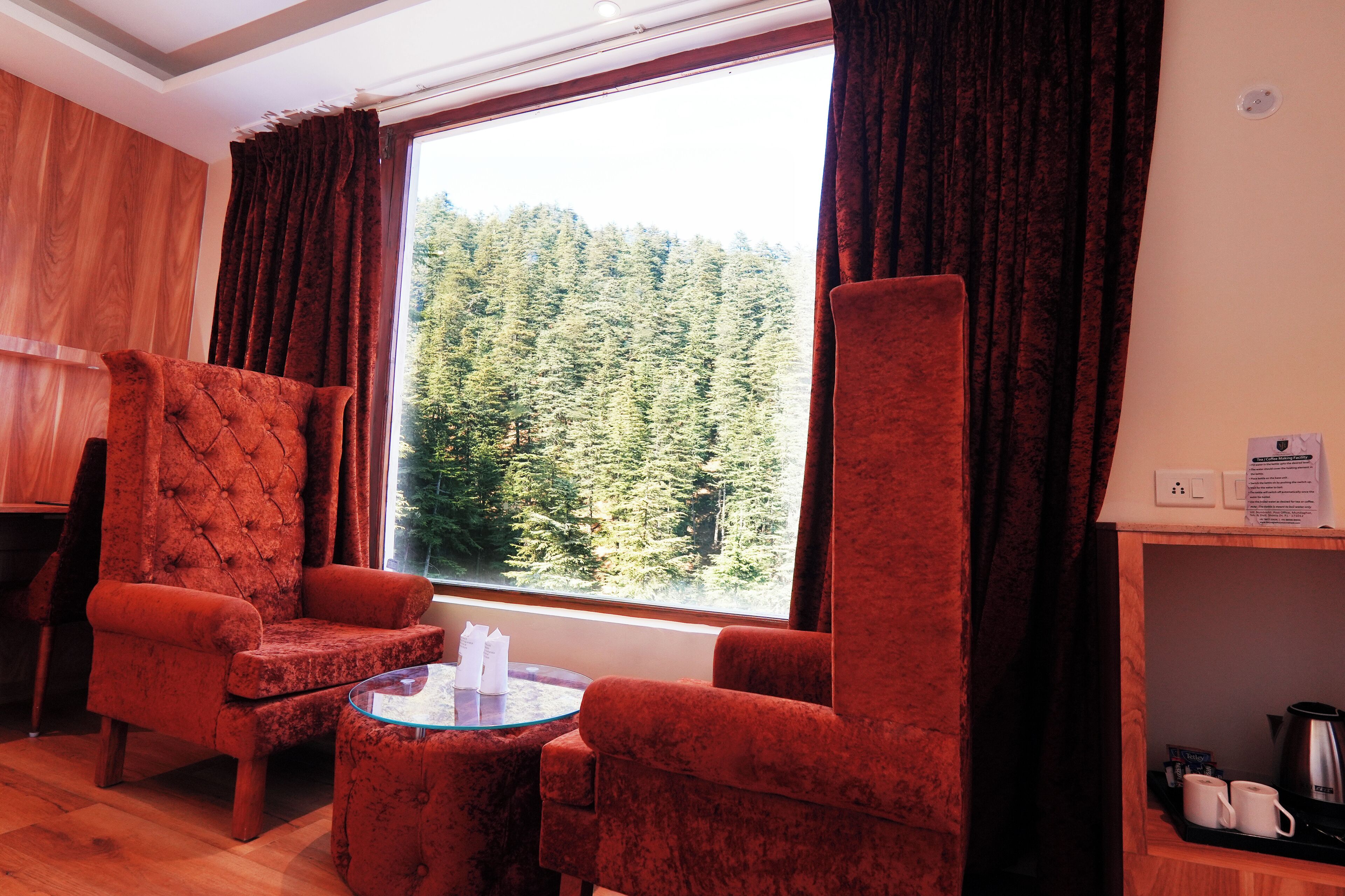 A room with a large window, two red armchairs, a small round table, and a wooden shelf with a kettle and cups.