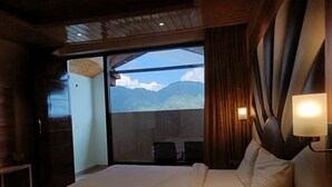 Habitación de hotel con una cama grande, vista a las montañas a través de una puerta corredera y un pequeño rincón de descanso con sillas.