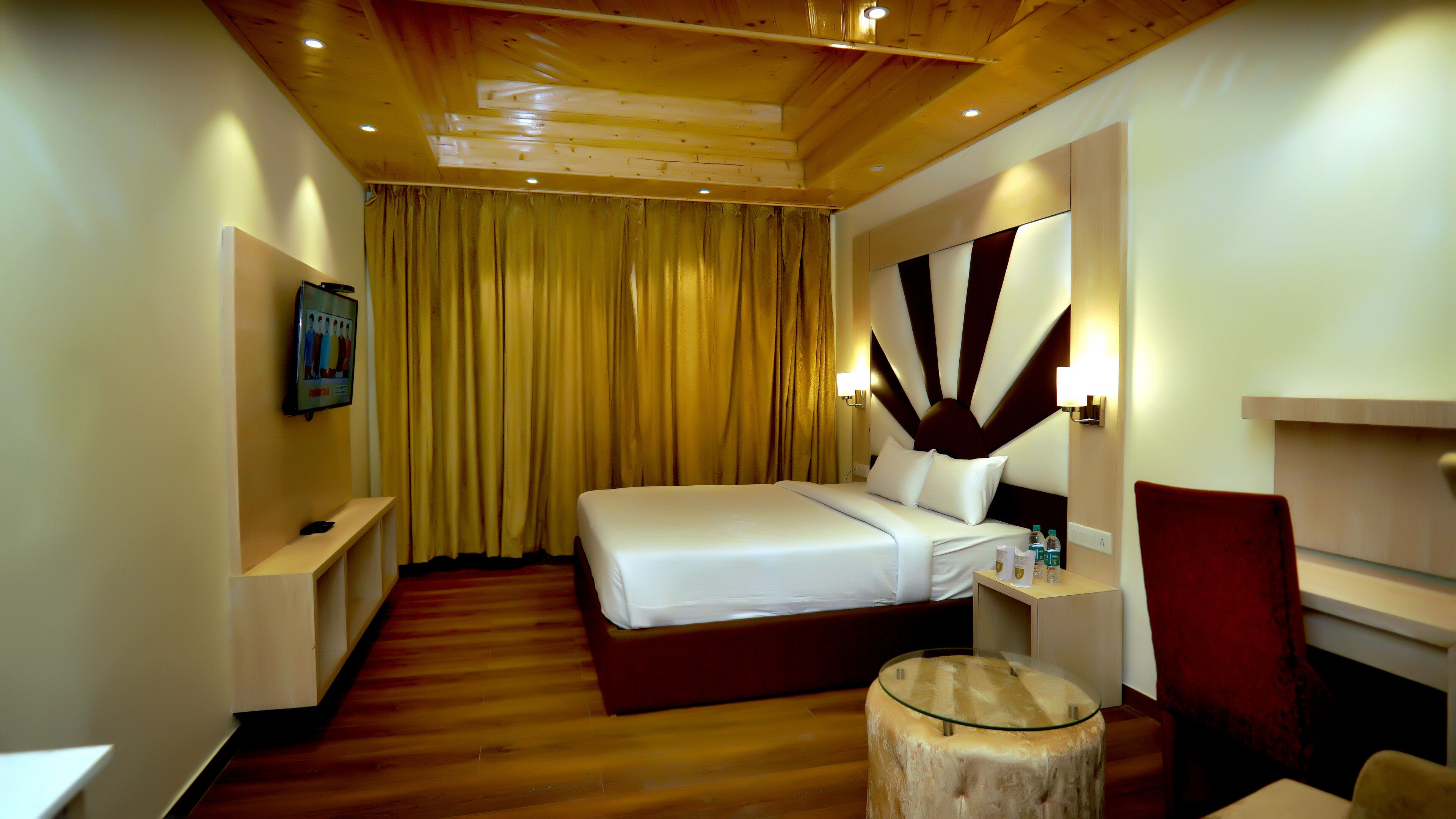 A hotel room with a bed, a bedside table, a small round table, a red chair, a mirror, and a curtain.