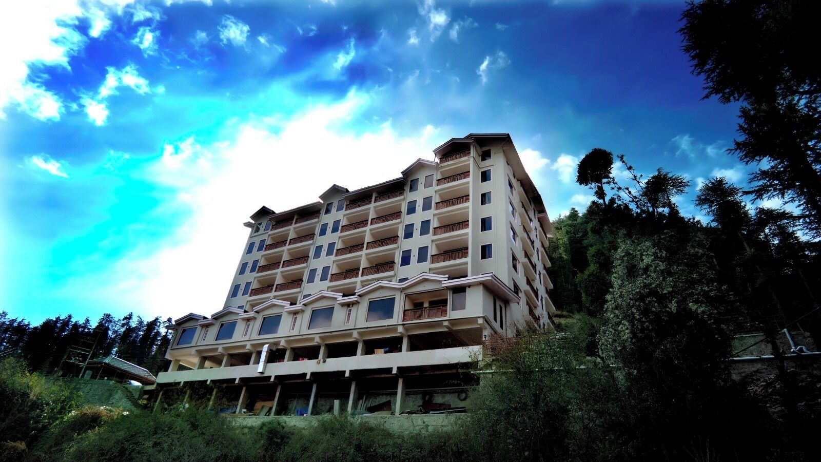 A multi-story building with balconies, surrounded by trees and under a partly cloudy sky.