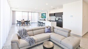 A modern living room with a sectional sofa, a dining area with a round table, and a kitchen with dark cabinets. - Indulge Apartments City View Penthouses (Mildura)