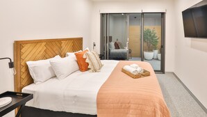 A hotel room with a wooden headboard, white bedding, an orange throw blanket, a bedside table with a lamp, and a TV mounted on the wall. - Indulge Apartments City View Penthouses (Mildura)