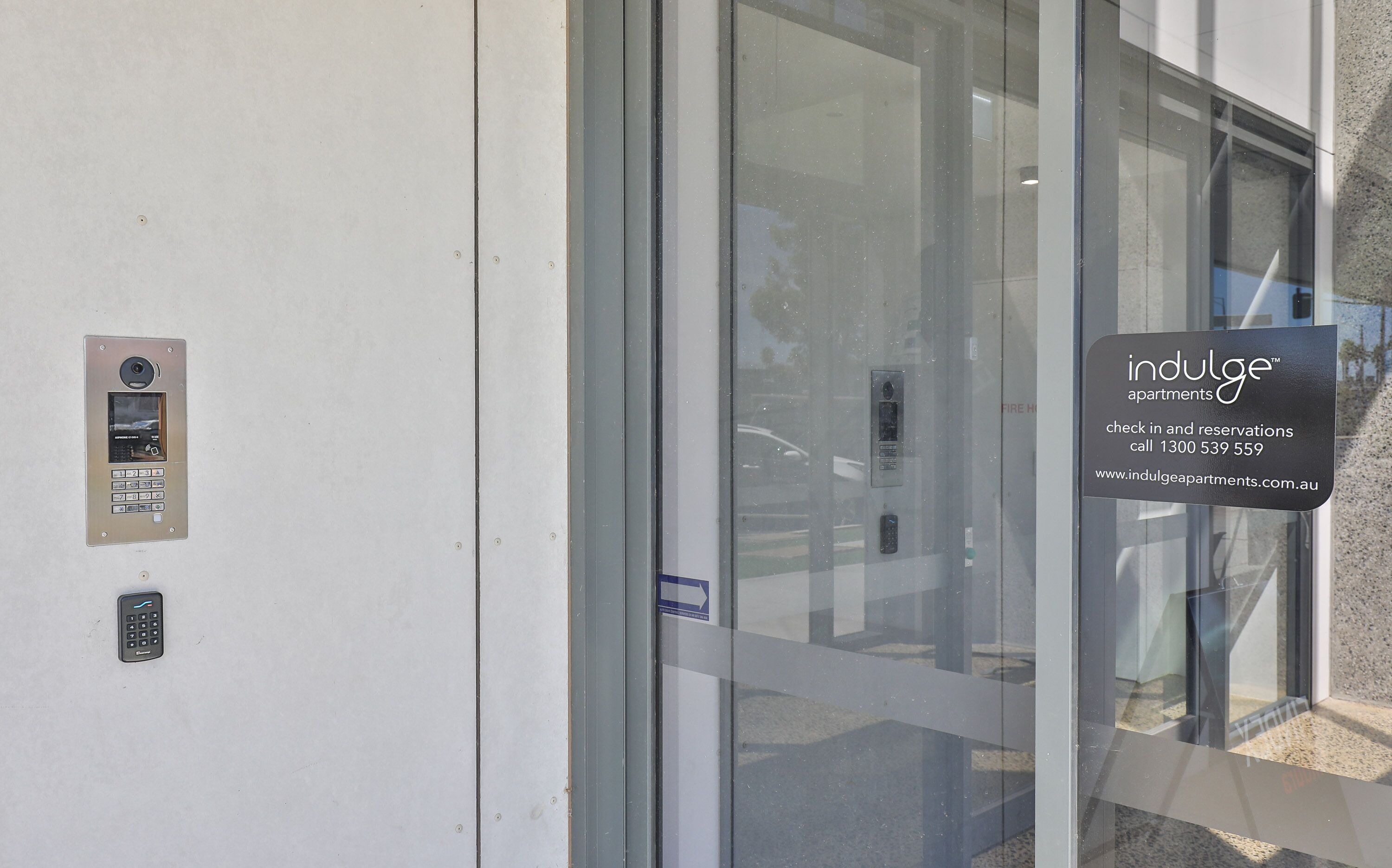 Ingresso moderno di un edificio con porta in vetro, tastierino per la sicurezza e cartello che indica Indulge Apartments con informazioni di contatto.