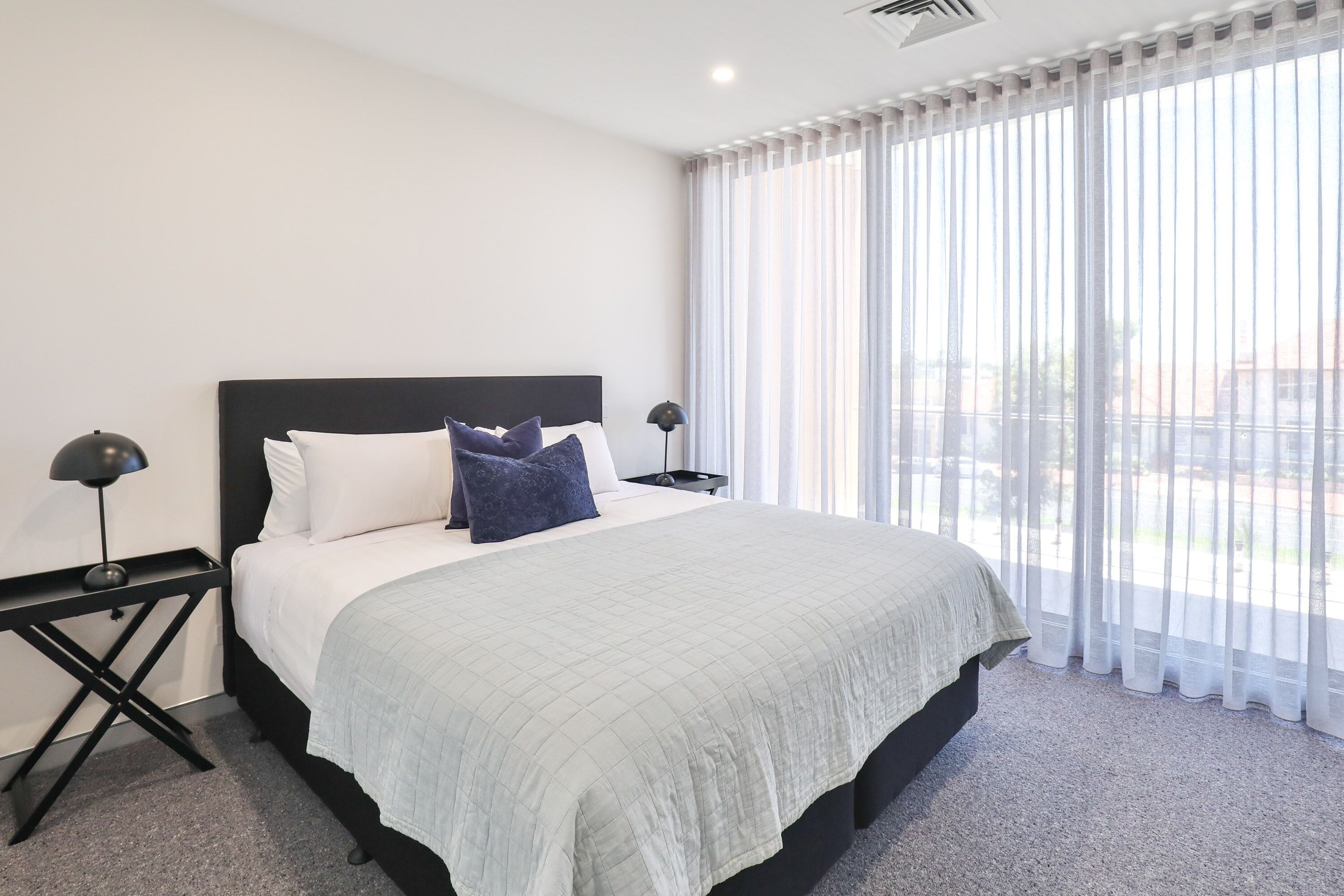 A modern hotel room with a large bed, two bedside tables, and a view through sheer curtains.
