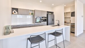 A modern kitchen with white countertops, stainless steel appliances, and black bar stools. - Indulge Apartments City View Penthouses (Mildura)