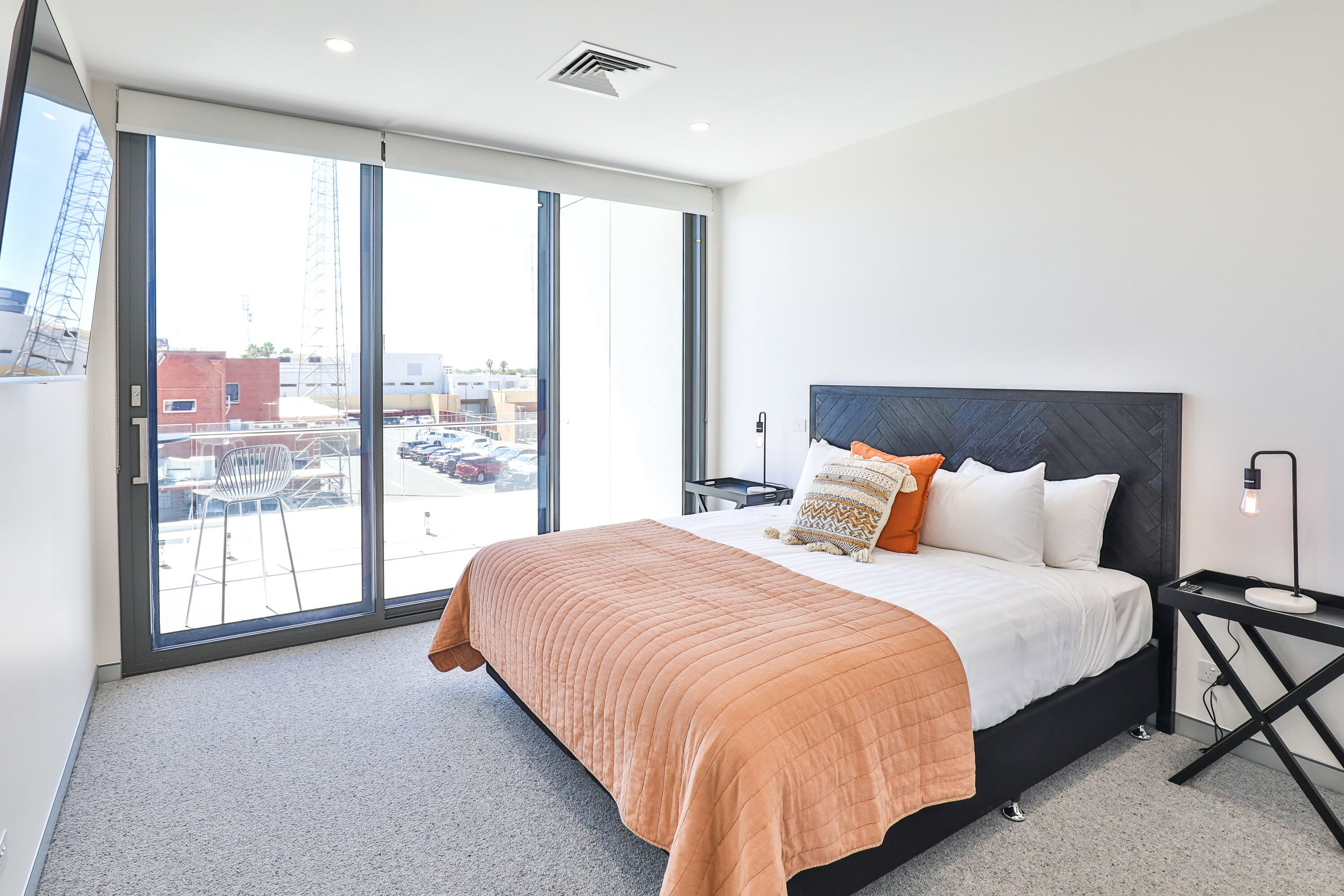 A modern hotel room with a large bed, a nightstand, a desk, and a view of the city through large windows.