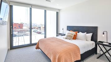 A modern hotel room with a large bed, a nightstand, a desk, and a view of the city through large windows.