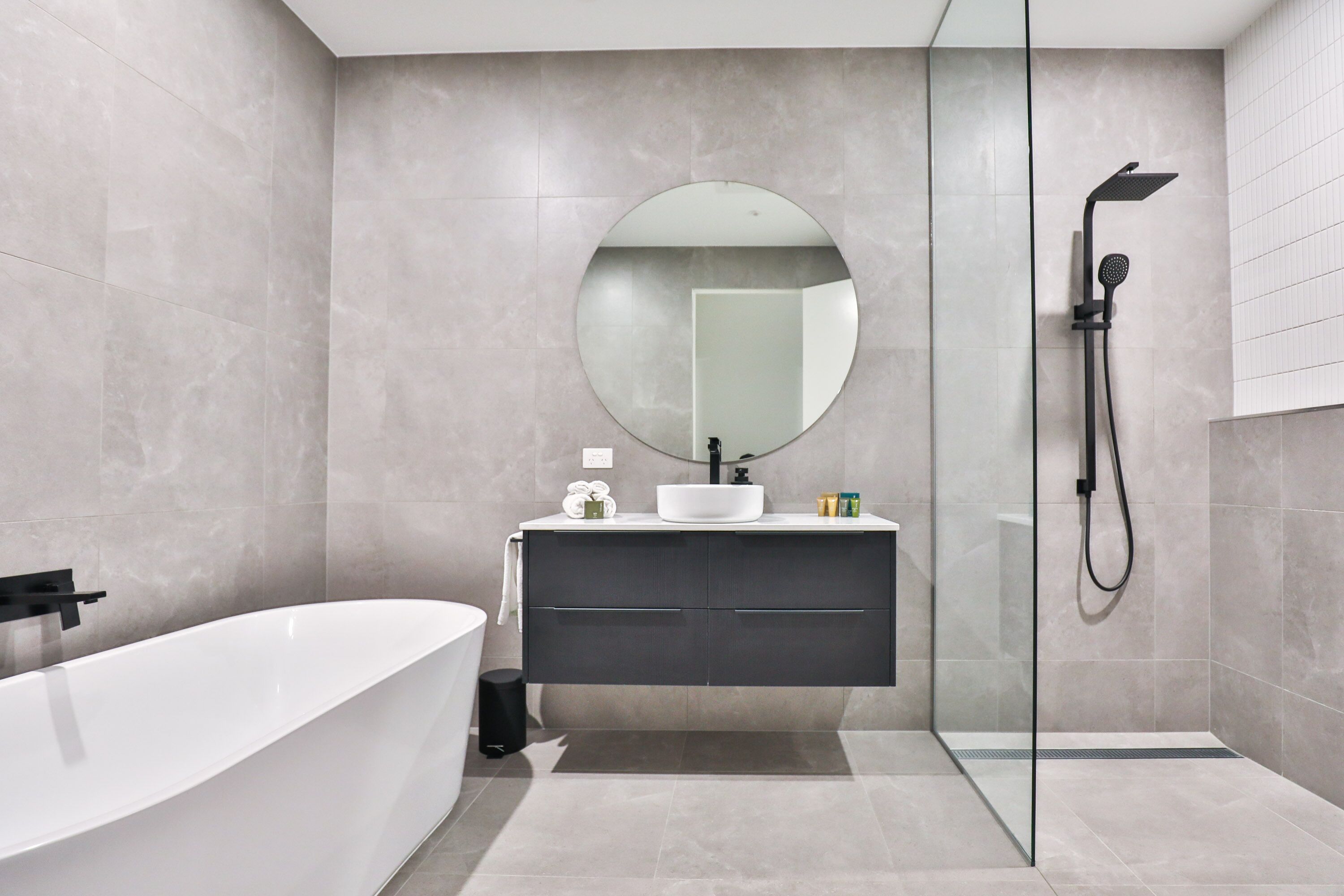 A modern bathroom with a freestanding bathtub, a round mirror, a glass-enclosed shower, and a dark-toned vanity.