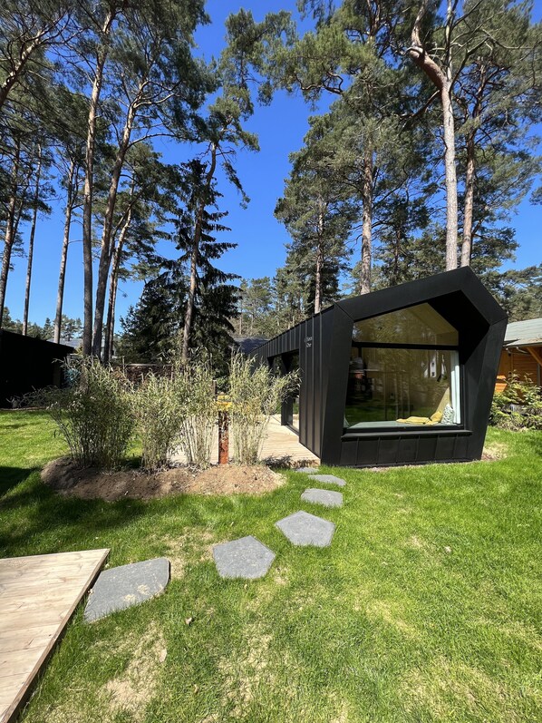 Une structure moderne, noire, entièrement vitrée, avec une terrasse en bois qui y mène, entourée d’une pelouse bien entretenue et de grands arbres.