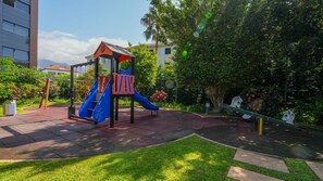 A playground with a blue slide and a red climbing wall, surrounded by greenery and a building in the background.