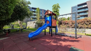 Un parque infantil con un tobogán azul y estructuras de escalada, rodeado de un área ajardinada con árboles y un muro de piedra.