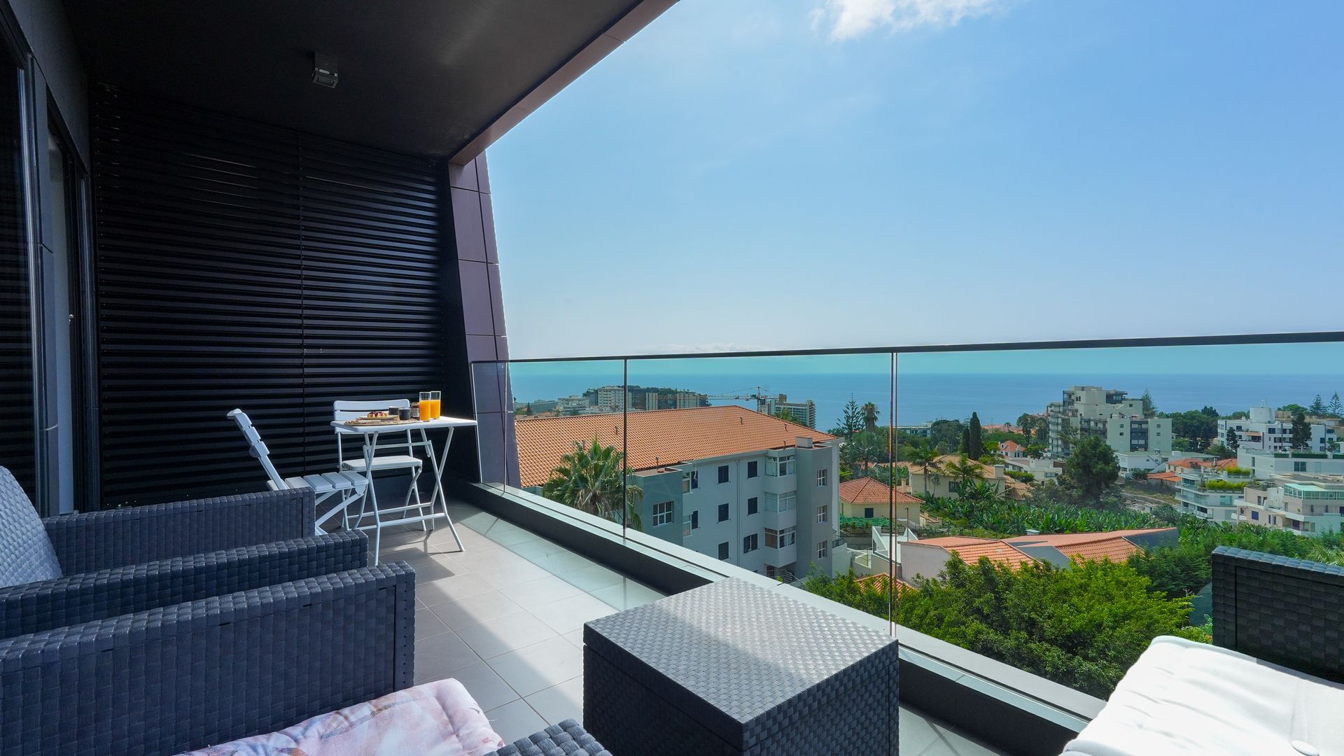 A balcony with wicker furniture, a table with a glass of orange juice, and a view of the sea and buildings.