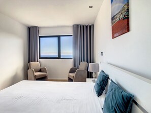 Quarto de hotel com uma cama grande, duas poltronas, uma mesa de cabeceira, uma luminária e uma janela com cortinas.