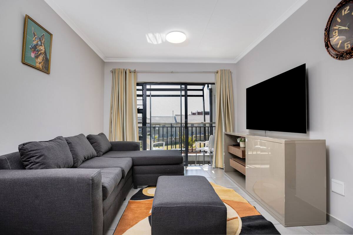 A modern living room with a grey sectional sofa, a flat-screen TV, and a balcony with curtains.