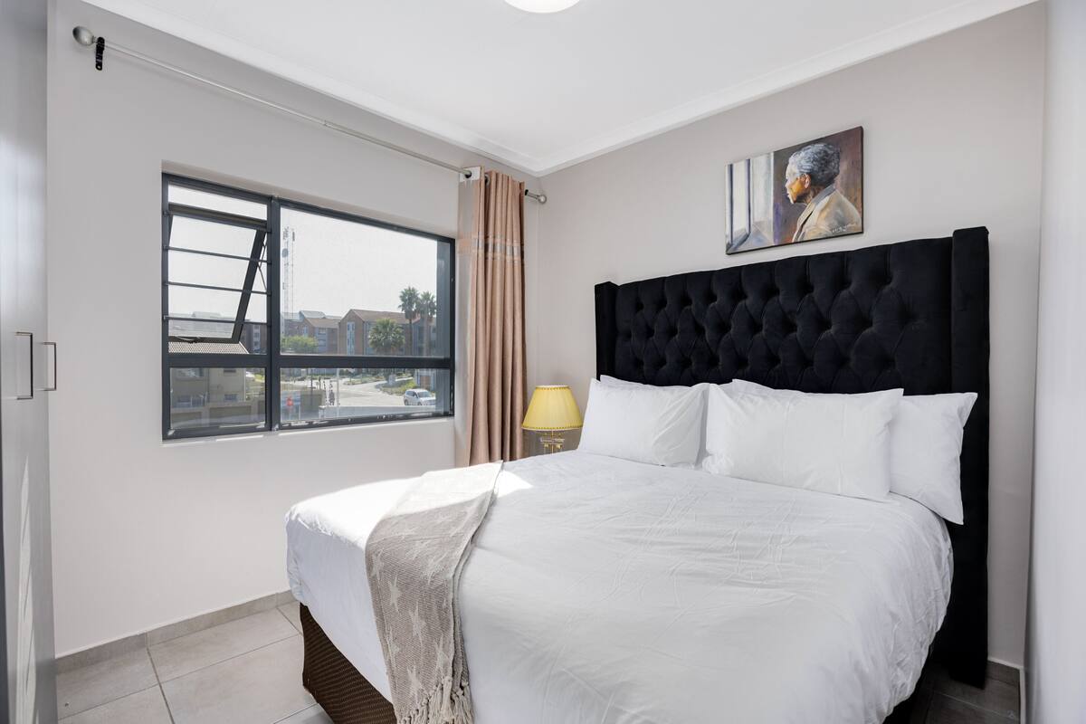 A hotel room with a large bed, a window with curtains, a lamp, and a painting on the wall.