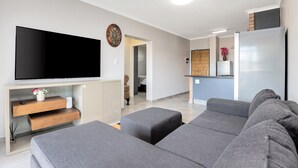 Una sala moderna con un televisor de pantalla plana, sofás grises, una mesa de centro de madera y una zona de cocina al fondo.