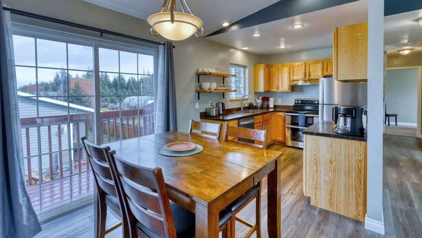 A modern kitchen with wooden cabinets, a dining area with a large table and chairs, and a view through large windows. - Adorable Alcove 2 Bedroom Escape Sleeps 4 (Kodiak)