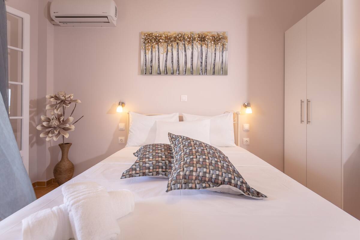 A neatly made bed with patterned pillows, a wall-mounted air conditioner, and a painting above it.