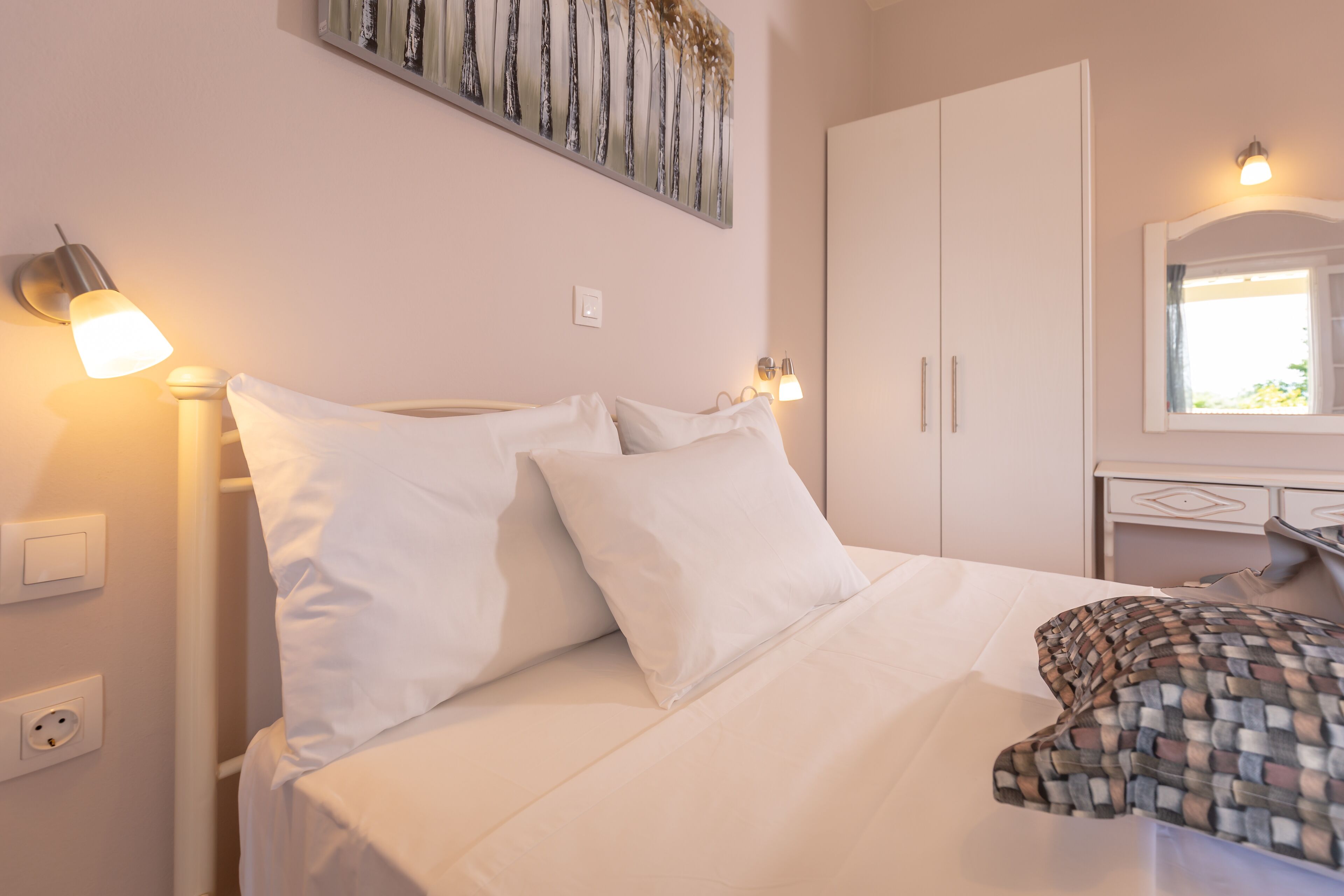 A neatly made bed with white bedding and pillows, a bedside lamp, a wall-mounted artwork, and a wardrobe with a mirror.