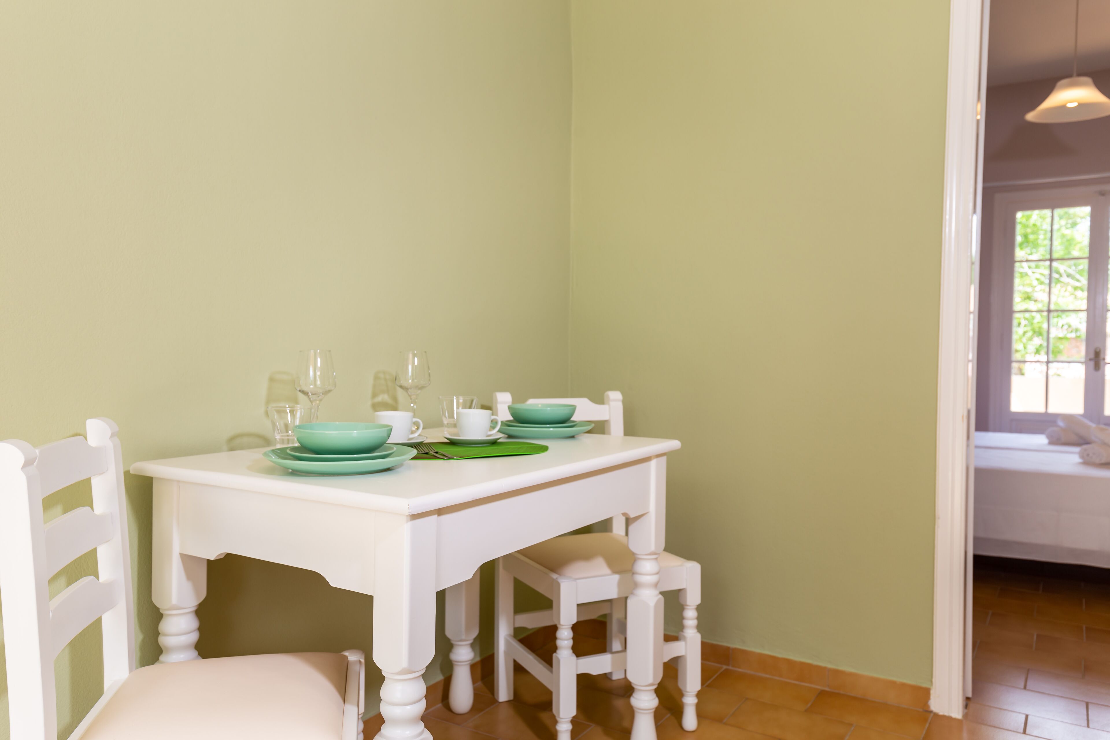 Un coin repas avec une table blanche dressée pour quatre personnes, comprenant des assiettes vertes et des verres transparents.