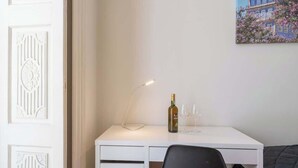 Un escritorio blanco con una botella y vasos, una silla negra y un cuadro de un edificio en la pared.