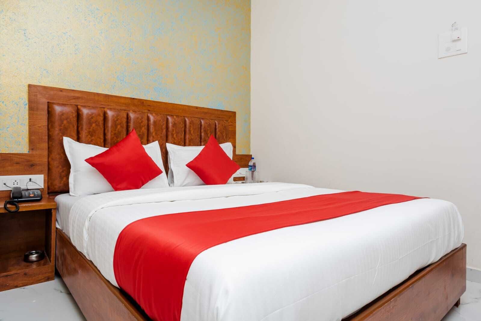 A neatly made bed with white and red bedding, a wooden headboard, and a wooden nightstand with a phone and a water bottle.
