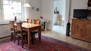 Un coin repas avec une table et des chaises en bois, un tapis à motifs, un radiateur, une fenêtre avec des rideaux et un téléviseur posé sur un meuble en bois.