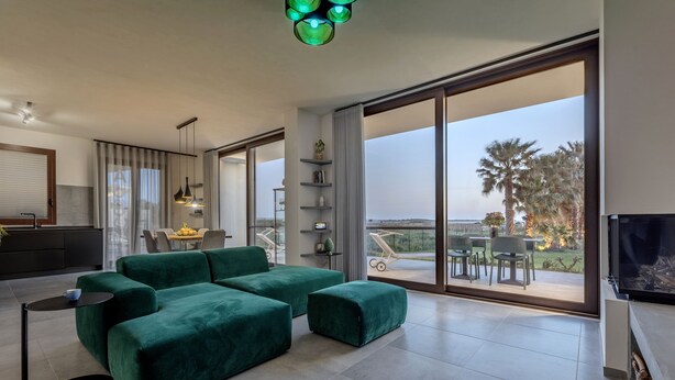 A modern living room with a green sectional sofa, a flat-screen TV, and a balcony with outdoor seating. - luxury holiday home in Lo Stagnone I Palm Kite Paradise I Casa A (Marsala)
